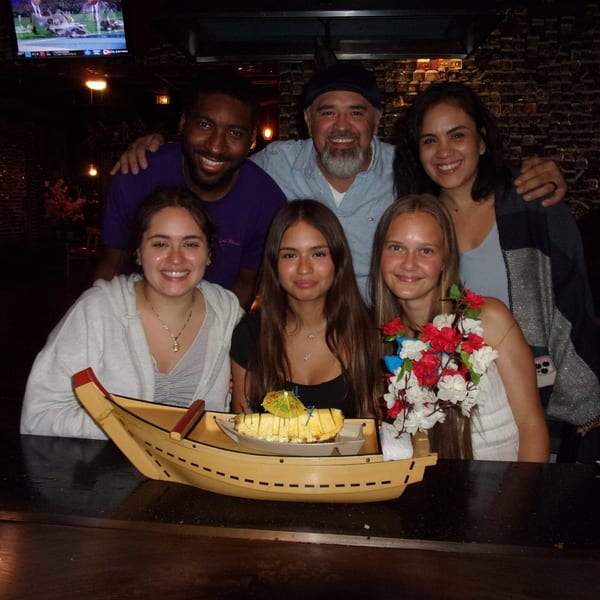 Six smiling people gathered around a hibachi table with a boat-shaped dish of pineapple.