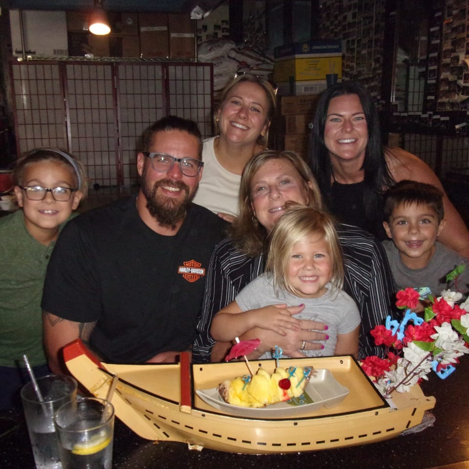 A family of seven smiles at Shogun Japanese Steakhouse, with a pineapple dessert boat in the foreground.