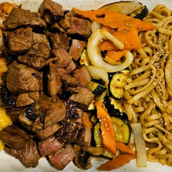 Hibachi steak, zucchini, carrots, onions, and noodles with sesame seeds on a white plate.