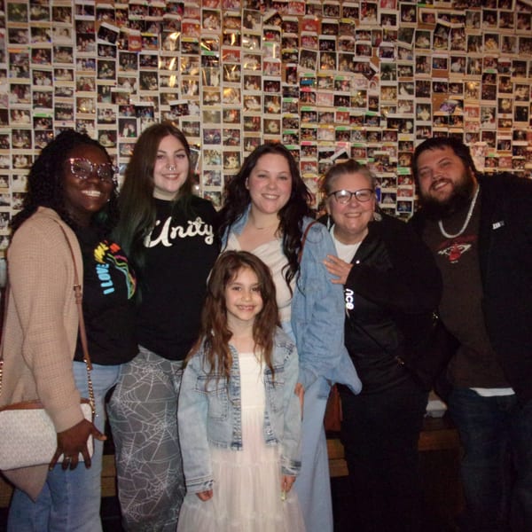 Six smiling people, including a child, pose in front of a wall covered in small photos.