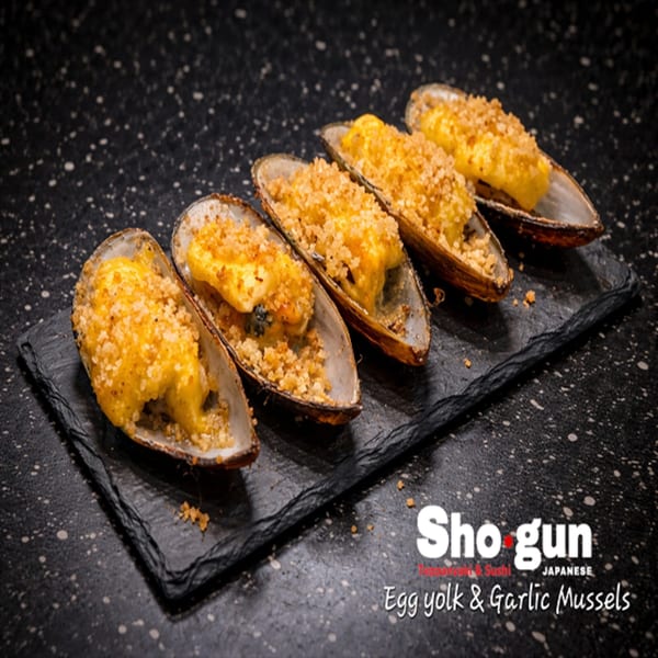 Five Shogun Japanese egg yolk and garlic mussels, topped with breadcrumbs, on a black slate.
