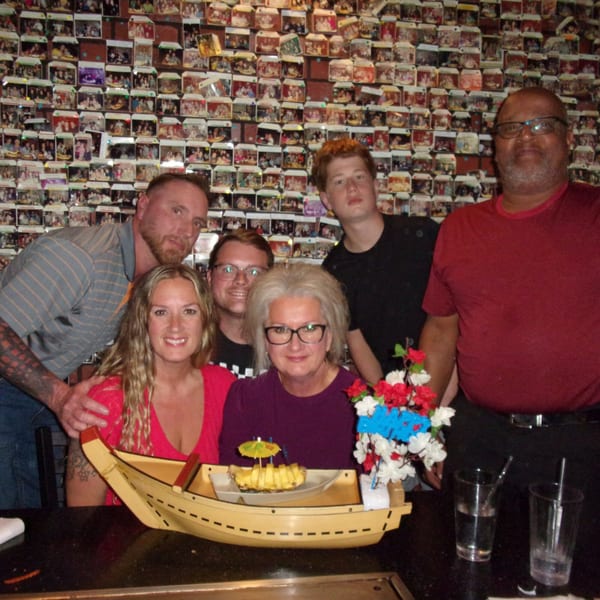 Six people pose for a photo at a hibachi table with a pineapple boat dessert and floral decor.