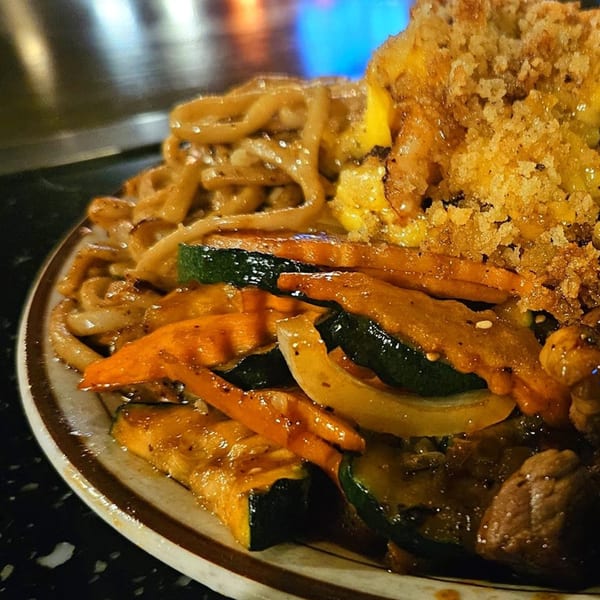 A hibachi plate with noodles, zucchini, carrots, and fried rice, with a blue flame in the background.